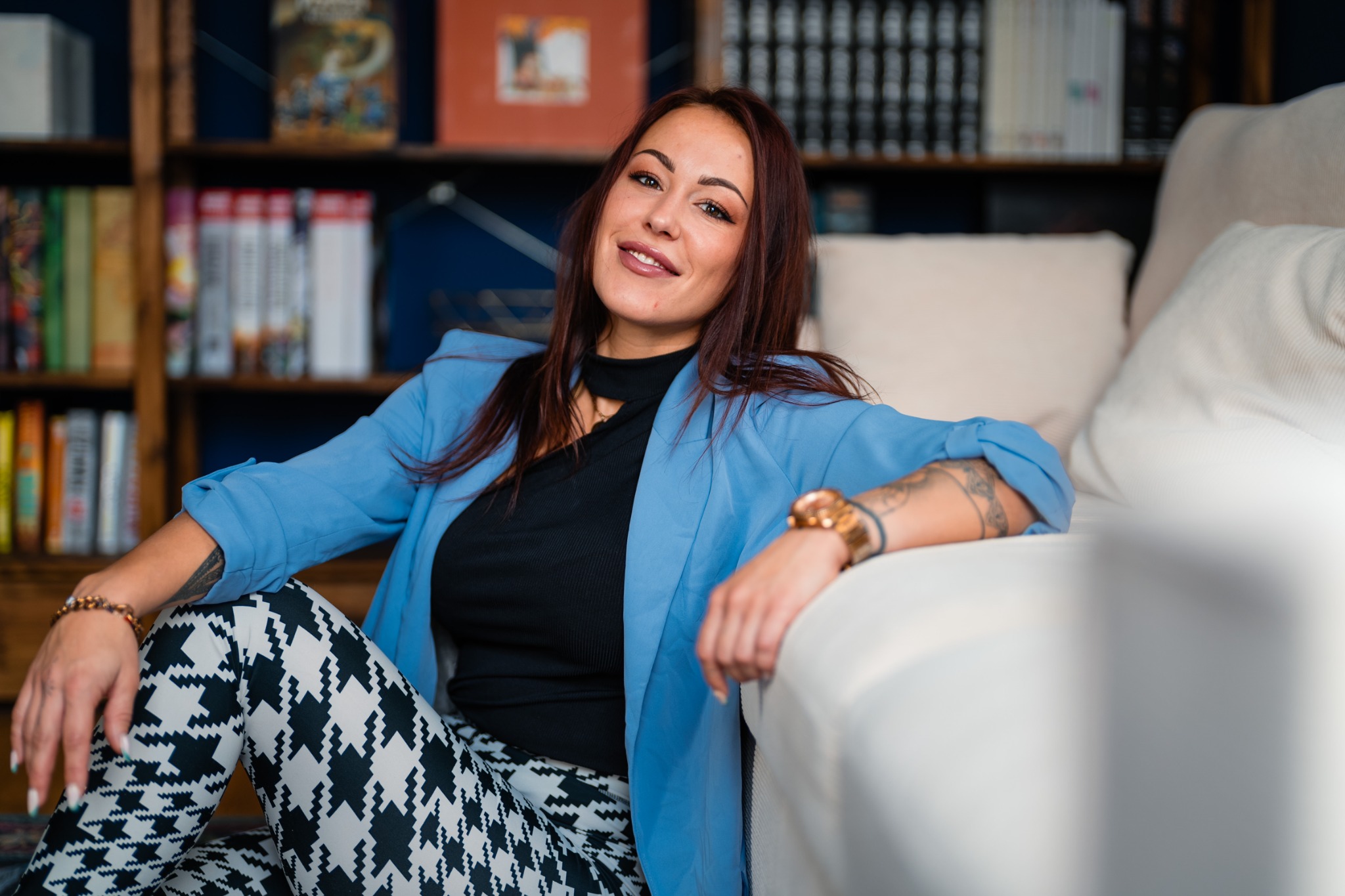 Lara sitzt entspannt im blauen Blazer vor einem Bücherregal